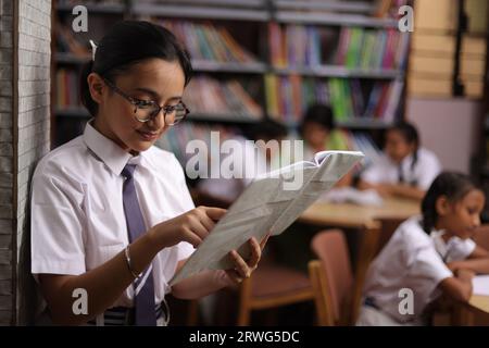 Smart student leaning against wall, reading a book in hand, focused, nerd, reader, in library. Stock Photo