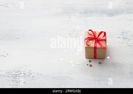 A small gift wrapped in kraft paper with a red ribbon on a light blue background with natural Christmas tree branches, surrounded by a shiny confetti Stock Photo
