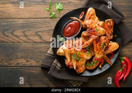 Chicken wings. Grilled or baked chicken wings with sesame seeds and ketchup or spicy tomato sauce on black plate on old wooden brawn table background. Stock Photo