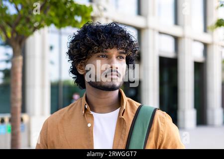 Close-up photo of a young Indian male programmer, designer, freelancer standing outside on the street with a backpack and seriously looking to the side. Stock Photo