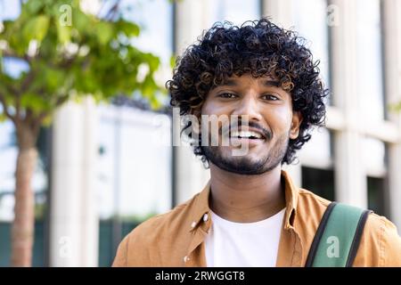 Close-up photo of a young Indian man wearing an orange shirt and carrying a backpack standing outside on the street and smiling at the camera. Stock Photo