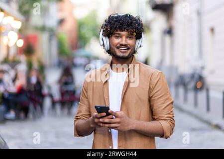 Portrait of a young handsome Indian man standing outside on the street wearing white headphones and holding a phone, smiling at the camera. Stock Photo