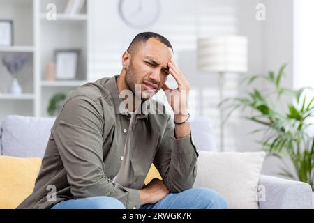 A young Muslim man is suffering from a headache. He sits on the sofa tiredly and massages his hand, grimacing from severe pain and tension. Stock Photo
