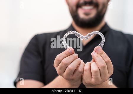 Smiling doctor holding heart made of clear aligners for professional teeth correction in clinic Stock Photo