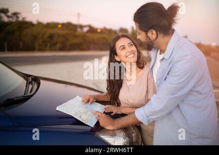 Enthusiastic latin couple reading map while having car trip, standing near the hood of car and talking, free space Stock Photo