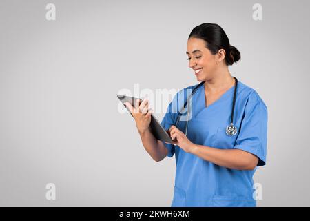 Glad latin lady nurse in blue uniform typing on tablet, working with technology, posing on grey background, free space Stock Photo