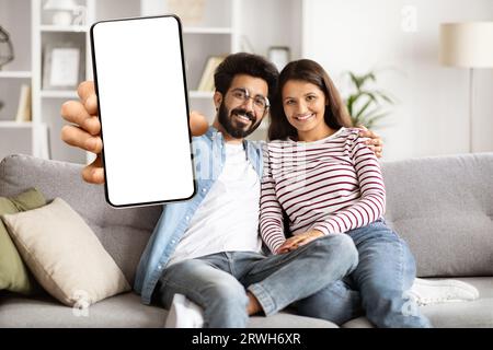 Cute happy indian couple sitting on couch, showing phone, mockup Stock Photo