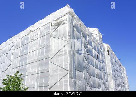 With white safety net covered multistorey residential building facade during renovation works, blue sky background Stock Photo