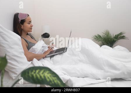 woman in blanket, multitasks by working online and studying remotely with laptop from her bed. Stock Photo