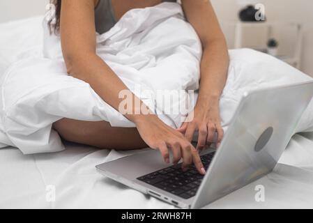 Focused on her laptop, a woman hands communicate in online social networks from comfort home bed. Stock Photo