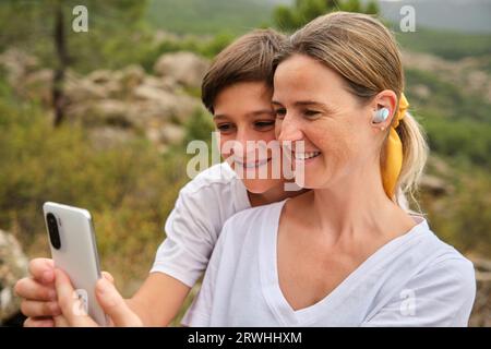 Happy adult mother and teenage son sitting with smartphone in daylight and attending video call Stock Photo