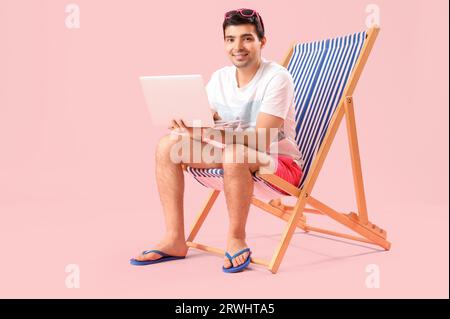 Young man with laptop sitting in deck chair on pink background Stock Photo