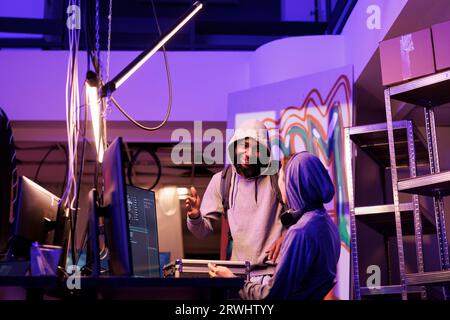 Men in hood discussing hacking plan with cheerful smile in abandoned warehouse. Happy young african american hacker explaining illegal ransomware code on computer to developer Stock Photo