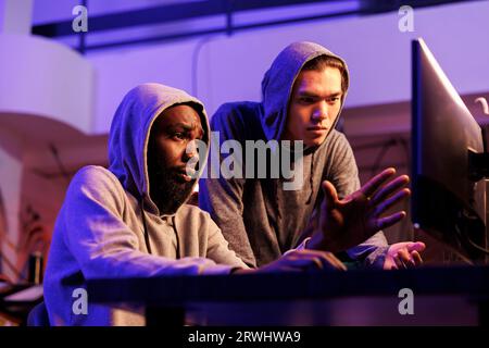 Criminals hacking server together and discussing password cracking malware. Confused african american and asian hackers in hoods checking malicious software on code on computer screen Stock Photo