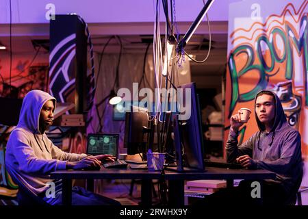 Hackers breaking into system and using spyware on computer while drinking coffee. Two criminals attacking website and programming internet virus malicious software at night Stock Photo