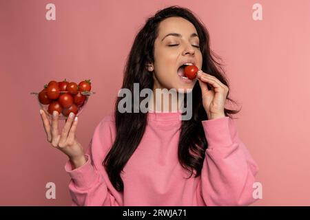 Hungry dark-haired female eating her favorite fresh vegetables with her eyes closed during the photo shoot. Raw food and healthy nutrition concept Stock Photo