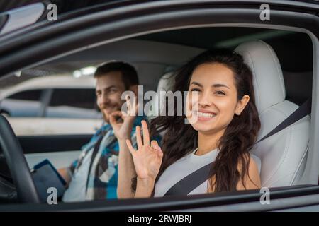 Happy woman and male instructor showing ok gesture while sitting in the car during drive test. Driving test, driver courses, exam concept Stock Photo