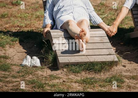 A couple of cute elderly people lie on sunbeds holding hands, basking in the sun. Detail shot, legs and arms. Stock Photo
