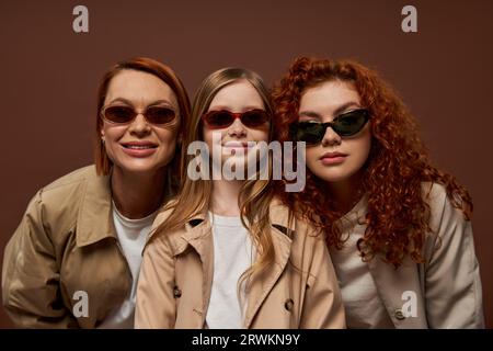 portrait of happy three generations of women looking at camera in sunglasses on brown backdrop Stock Photo