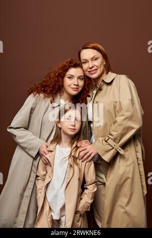 three generations, cheerful women and girl posing in autumn trench coats on grey background, fashion Stock Photo