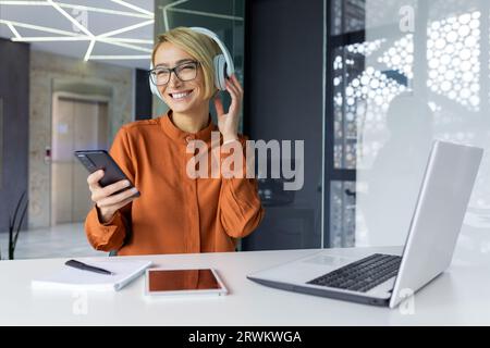 Businesswoman at workplace listening to music in headphones, using app on phone, female worker singing along joyfully and dancing, holding smartphone while sitting at table with laptop. Stock Photo