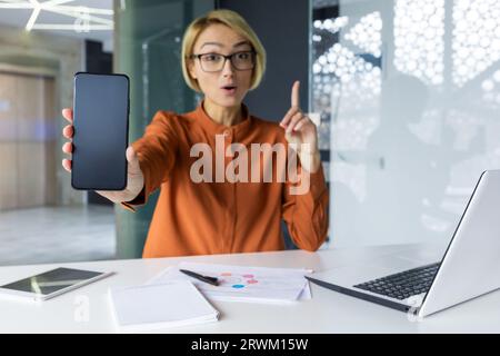 Joyful and successful business woman shows phone screen to camera, blank space for recording. Woman at workplace recommending financial app using smartphone inside office. Stock Photo