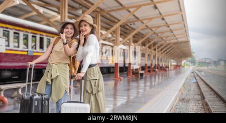 Two women are happy while traveling at the train station. tour concept. Two asian women best friend travel pointing finger on train timetable in Stock Photo