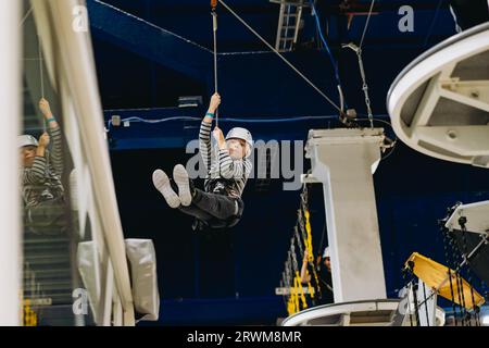 caucasian boy of 8 years old climbing in adventure park passing obstacle course going down on zipline. high rope park indoors . Stock Photo