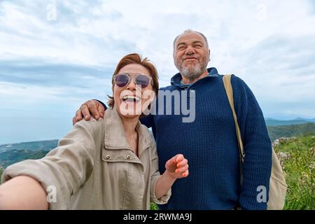 Middle aged traveler couple have fun in mountain using smartphone to take selfie. Mental health, wellbeing, trip adventure and healthy lifestyle. Stock Photo