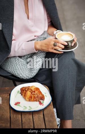 Girl is sitting in cafe and holding cup of cappuccino coffee in her hands, comfortably wrapped in gray plaid scarf. Close up of female hands. Stock Photo
