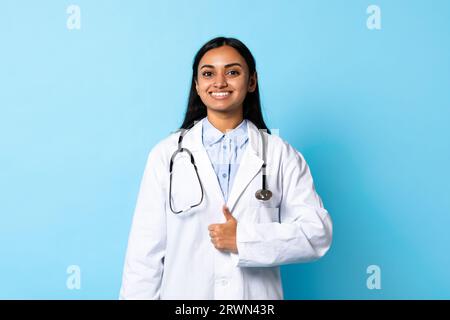 Smiling Indian Doctor Young Woman Gesturing Thumbs Up, Blue Background Stock Photo