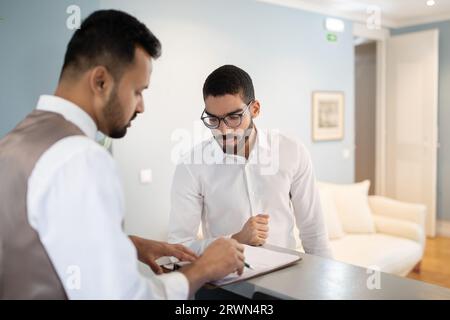 Arabic Businessman Checking In At Hotel, Signing Papers With Receptionist Stock Photo