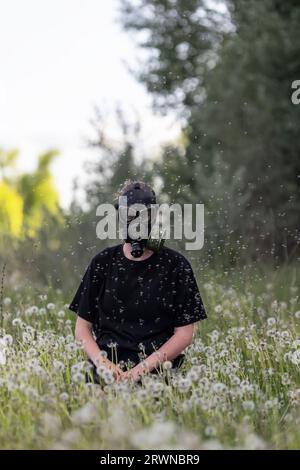 girl in a gas mask protects herself from allergies from flying dandelion seeds sitting in a spring meadow Stock Photo
