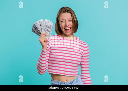 Rich pleased boss young woman waving money dollar cash banknotes bills like a fan, success business career, lottery winner, big income, wealth. Girl in crop top isolated on blue background, indoors Stock Photo