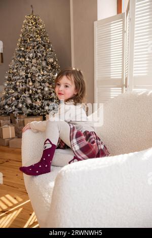 Funny little girl lies in a soft white chair, fooling around, making faces near the Christmas tree. Child in a cozy atmosphere at home. Festive interi Stock Photo