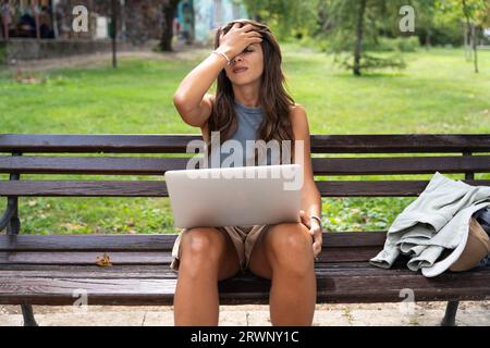 Young hipster woman with laptop facepalm gesture, feeling regret, sorrow, blaming herself for mistake, raise her hand, sitting and working in park. Fr Stock Photo