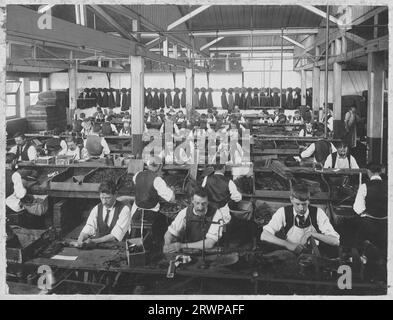 The Cigar making room at States Tobacco Company, A'Beckett Street, Melbourne. Elevated view of interior showing rows of benches, men in shirtsleeves and aprons, making cigars, bundles of cigars on bench in front of them and on table lower left. Young apprentices visible in some areas. Hand rolling. Row of coats and hats visible on back wall. Image dated between 1900 and 1920 Stock Photo