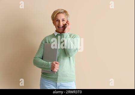 Mature female programmer with laptop talking by mobile phone on beige background Stock Photo