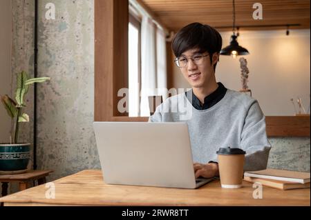 A happy and smart young Asian male college student in casual clothes and eyeglasses is using his laptop computer while sitting in a coffee shop. Stock Photo