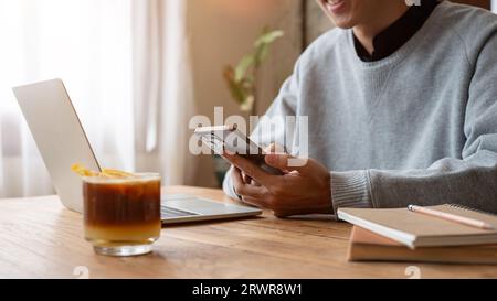 A happy Asian man in a sweater enjoys chatting with his friends on his phone, using his smartphone while working remotely at a coffee shop. Lifestyle Stock Photo