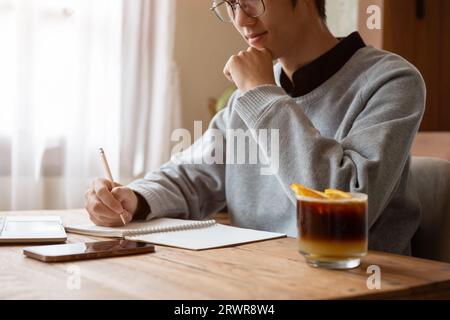 A smart and thoughtful young Asian male college student focuses on his homework, writing an essay or taking notes on his schoolbook, sitting in a cafe Stock Photo