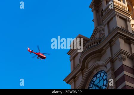 Red helicopter in flight with blue sky background near heritage clocktower building in Melbourne city, Australia. Stock Photo