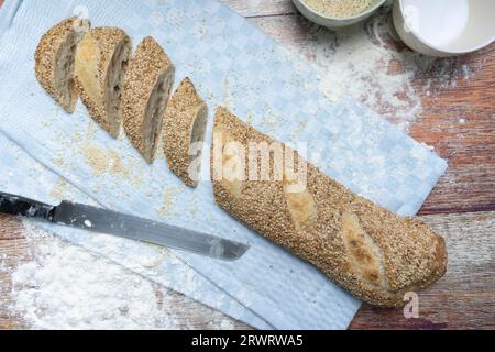 Freshly baked, vegan baguette with sesame seeds, cut on a light blue cloth, flour and sesame seeds around it, bread knife, two small bowls in cut, on Stock Photo