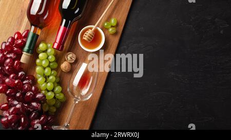 Red and white wine bottles with bunch of grapes, cheese, honey, nuts and wineglass on wooden board and black background. Top view with copy space. Stock Photo