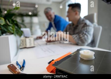 Two men architects using 3D printer in office and projecting house, drawing blueprints. Stock Photo