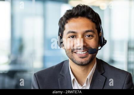 Close-up portrait of young successful businessman with headset phone, customer support worker in business suit smiling and looking at camera, workplace inside office. Stock Photo