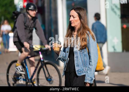 Young to middle aged lady walking across a road carrying a cardboard cup of coffee or another hot beverage, with cyclist in the background. Stock Photo