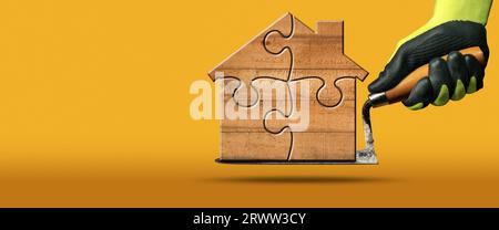 Manual worker with protective work gloves holding a dirty trowel and a small wooden house formed from jigsaw puzzle pieces. Stock Photo
