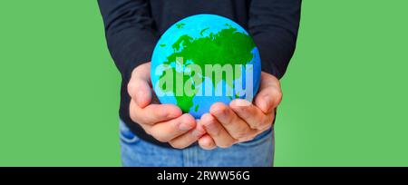 Person wearing black long sleeve and blue jeans extends hands to offer a verdant model of the Earth isolated on green background. Stock Photo