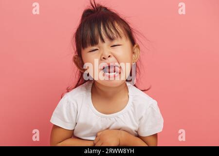 Stubborn three year old Asian little girl capriciously shows her tongue to the camera on a pink isolated background. Naughty child concept Stock Photo